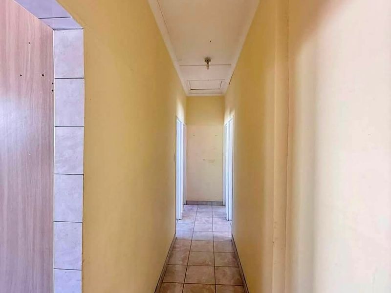 A narrow, empty residential hallway featuring tiled flooring and a ceiling access hatch, finished with plain yellow wall paint.