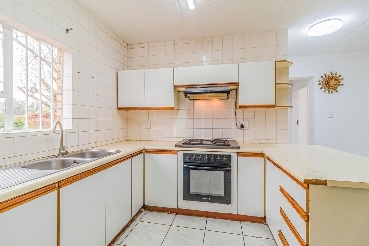 A dated kitchen featuring white cabinetry, built-in oven and hob, tiled splashbacks, and a double sink with security bars on the window.
