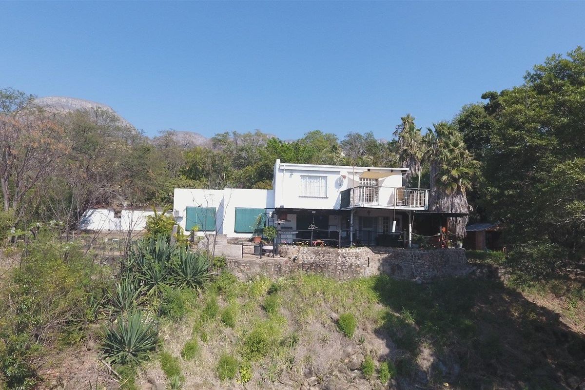 Elevated rear view of the farmhouse featuring a stone retaining wall, covered patio area, and an upper-level balcony set against a natural landscape.