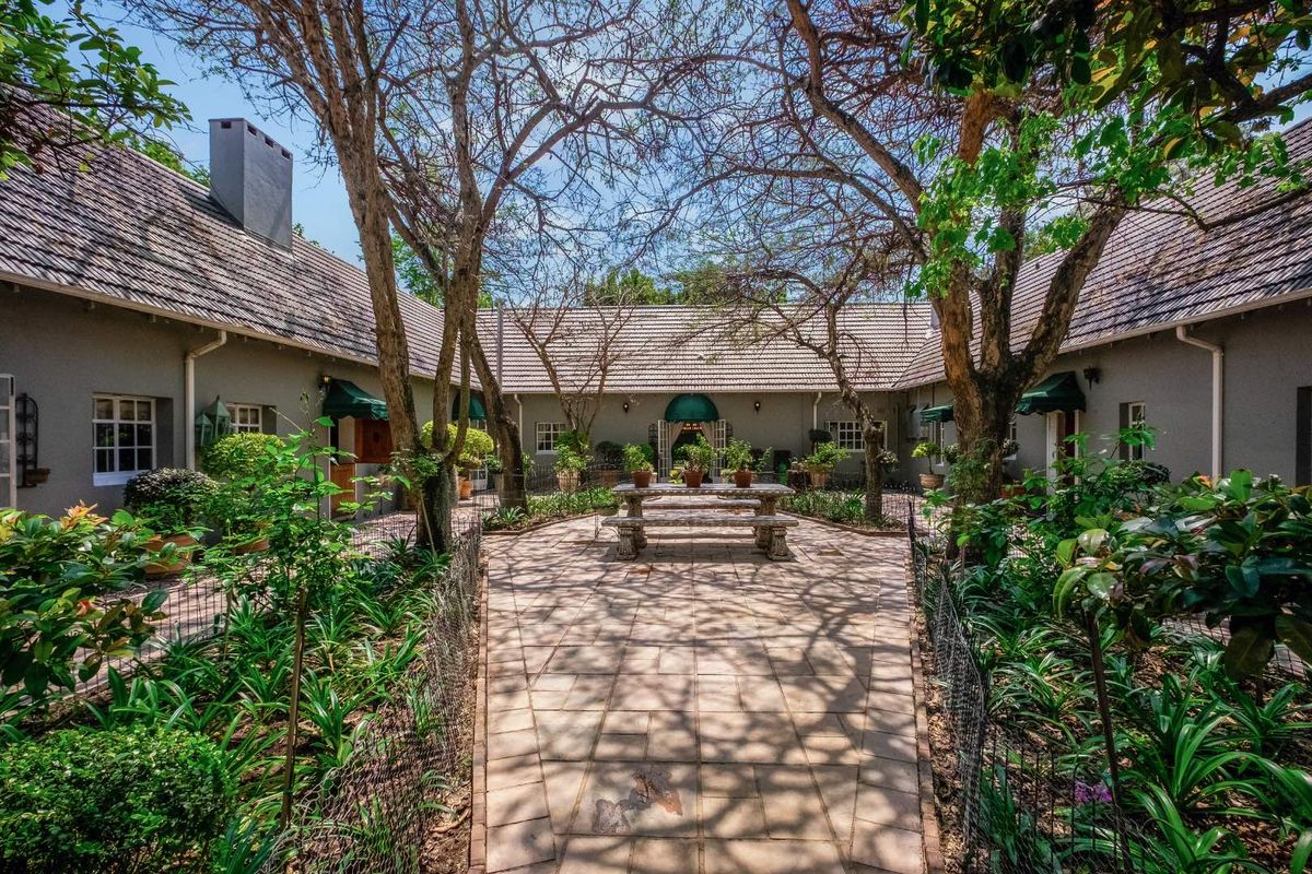 A charming U-shaped courtyard featuring a paved patio area, mature trees, and well-maintained garden beds surrounded by the home's exterior structure.