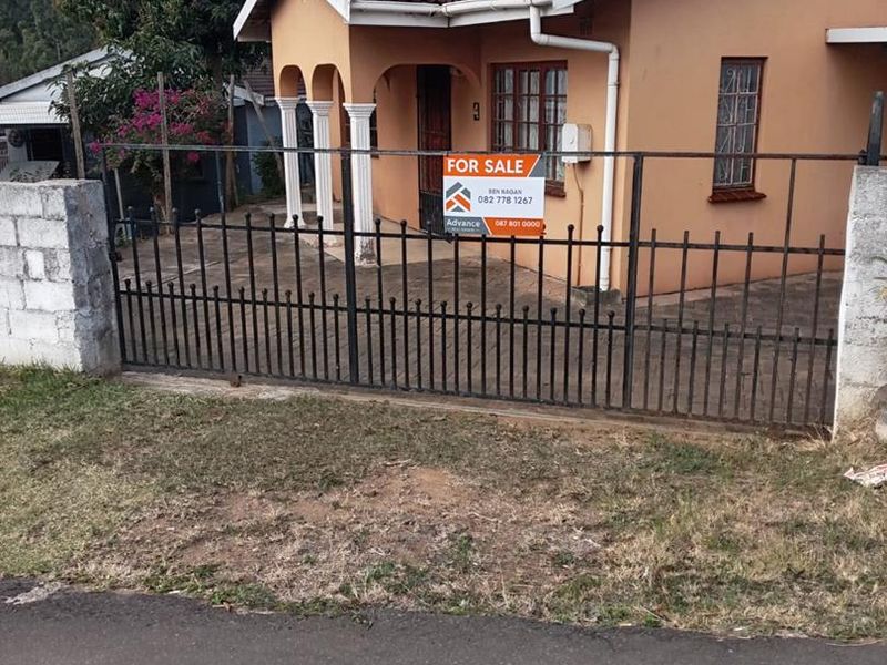 Front elevation of a single-story house featuring a tiled roof, decorative pillars, and a secure metal gate with a paved driveway.