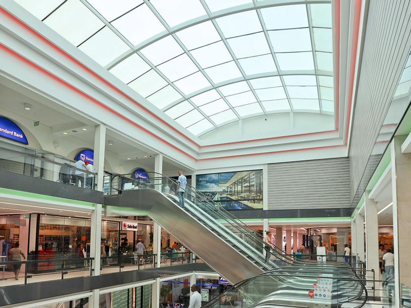 Modern multi-level retail shopping center interior featuring a large glass atrium skylight, central escalator, and contemporary storefronts.