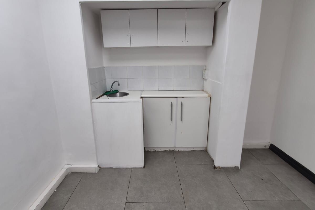 A compact kitchenette area featuring built-in white cabinetry, a stainless steel sink, tiled backsplash, and grey floor tiles in an office setting.