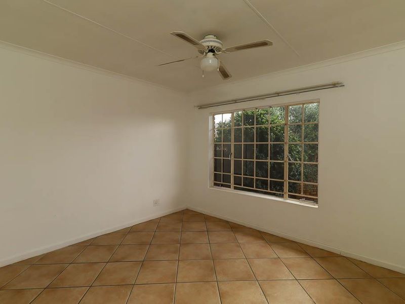 A bright, empty bedroom featuring neutral tiled flooring, a ceiling fan, and a large grid-pattern window overlooking the garden.