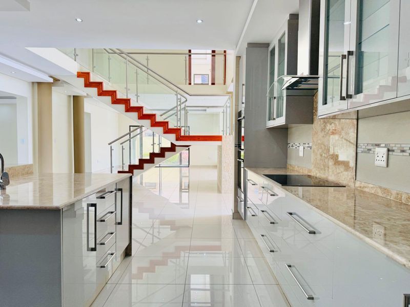 Modern open-plan kitchen featuring high-gloss cabinetry, stone countertops, integrated appliances, and a striking staircase with glass balustrades in a bright, tiled space.