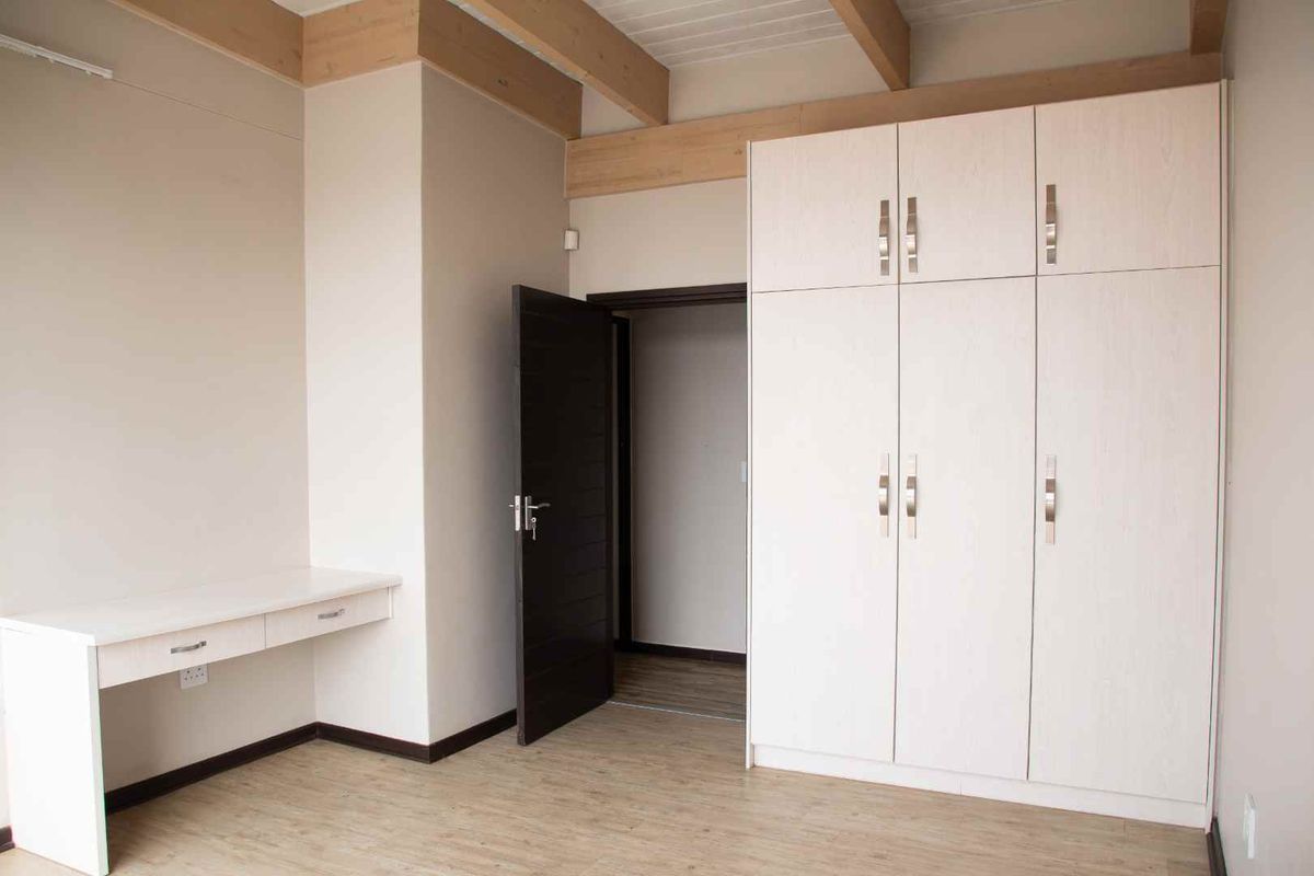A bright bedroom featuring exposed timber ceiling beams, light laminate flooring, a built-in desk, and ample storage with a large built-in wardrobe.