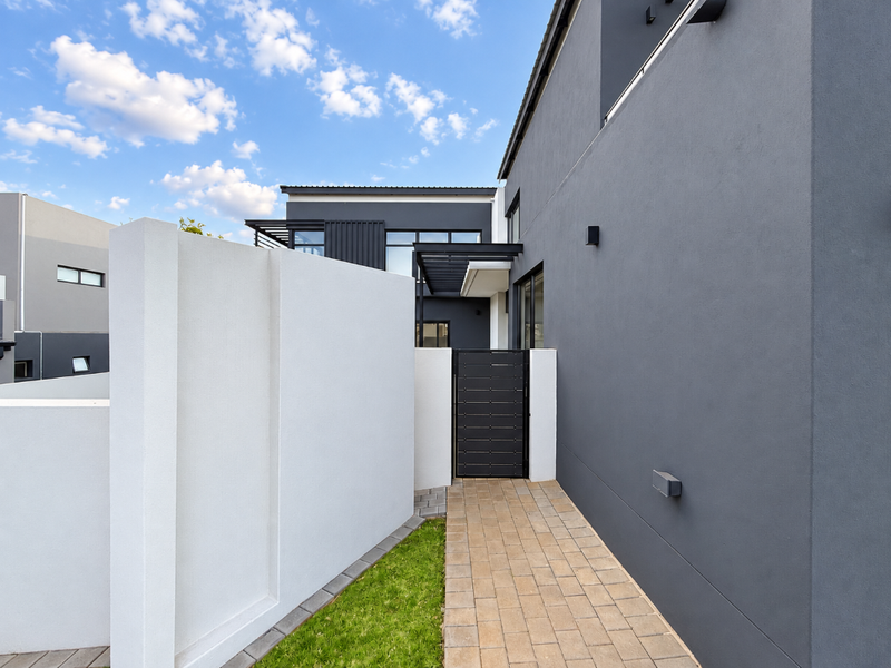 A modern rear exterior view featuring a paved walkway, a secure metal pedestrian gate, and clean rendered walls with integrated exterior lighting.