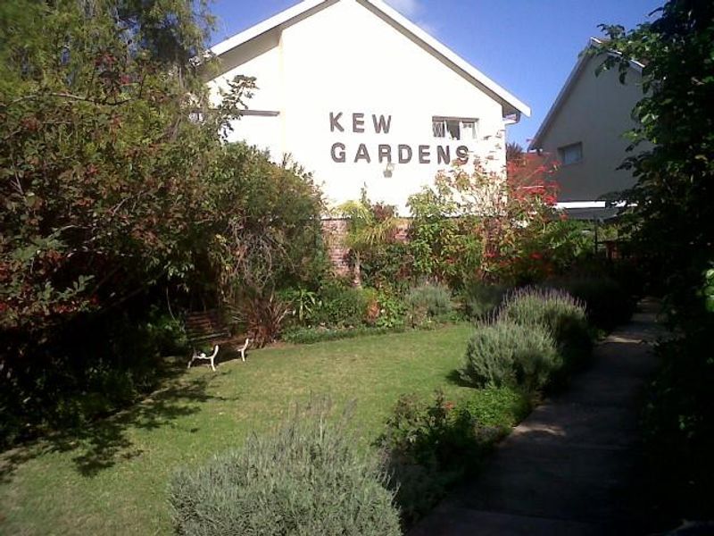 Front exterior view of the Kew Gardens apartment complex featuring a landscaped garden and paved walkway leading to the building entrance.