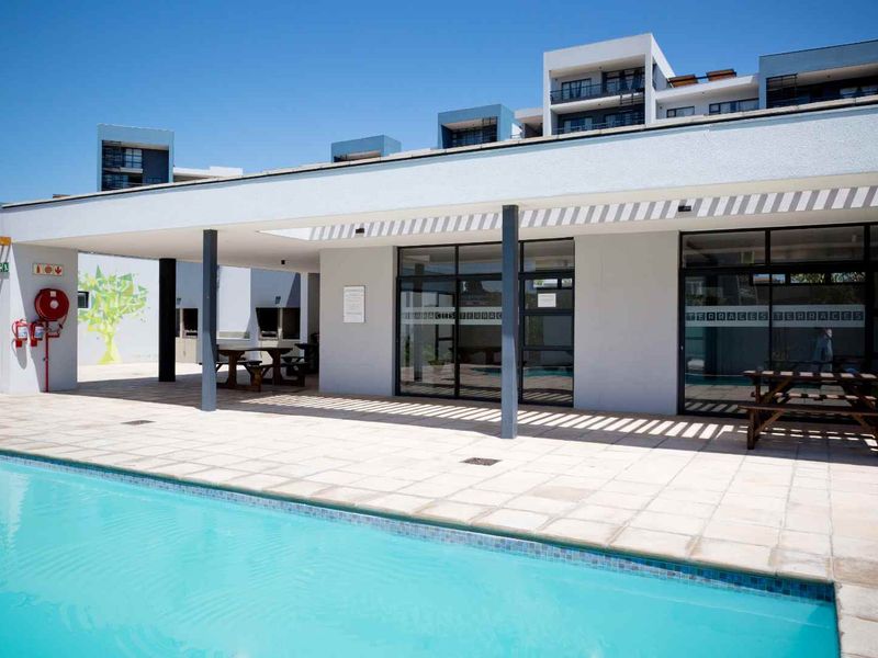 A well-maintained communal pool area featuring a tiled deck, covered patio space with built-in braai facilities, and modern glass access doors.