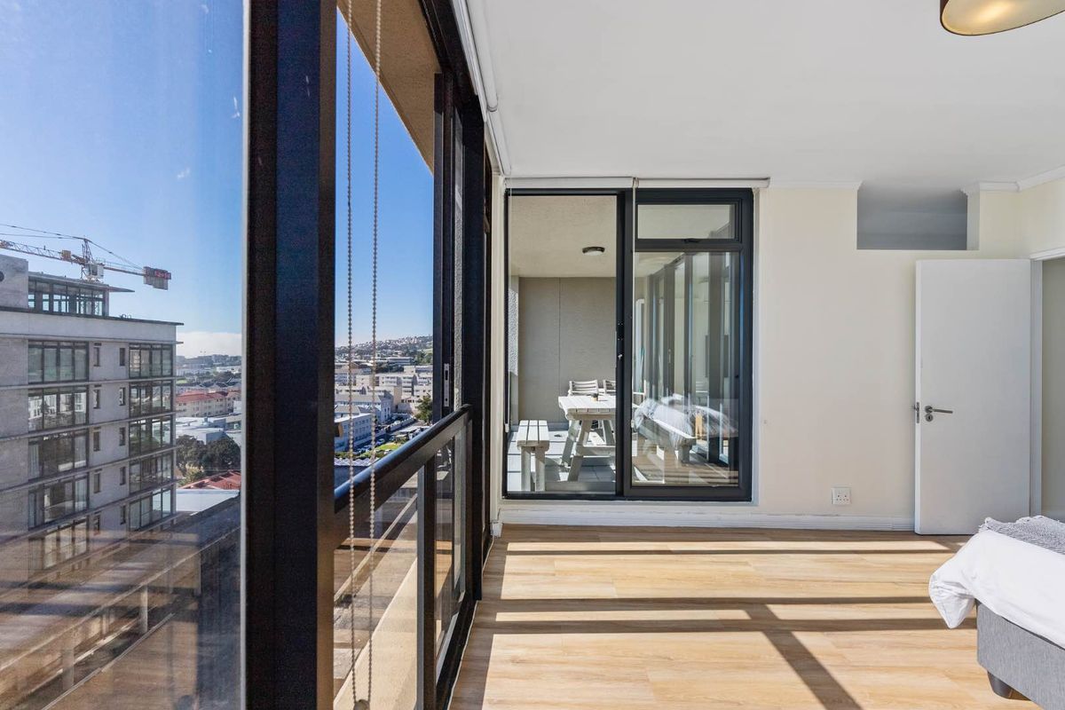 Bright bedroom featuring floor-to-ceiling glass sliding doors that open onto a private balcony with expansive city views and modern laminate flooring.