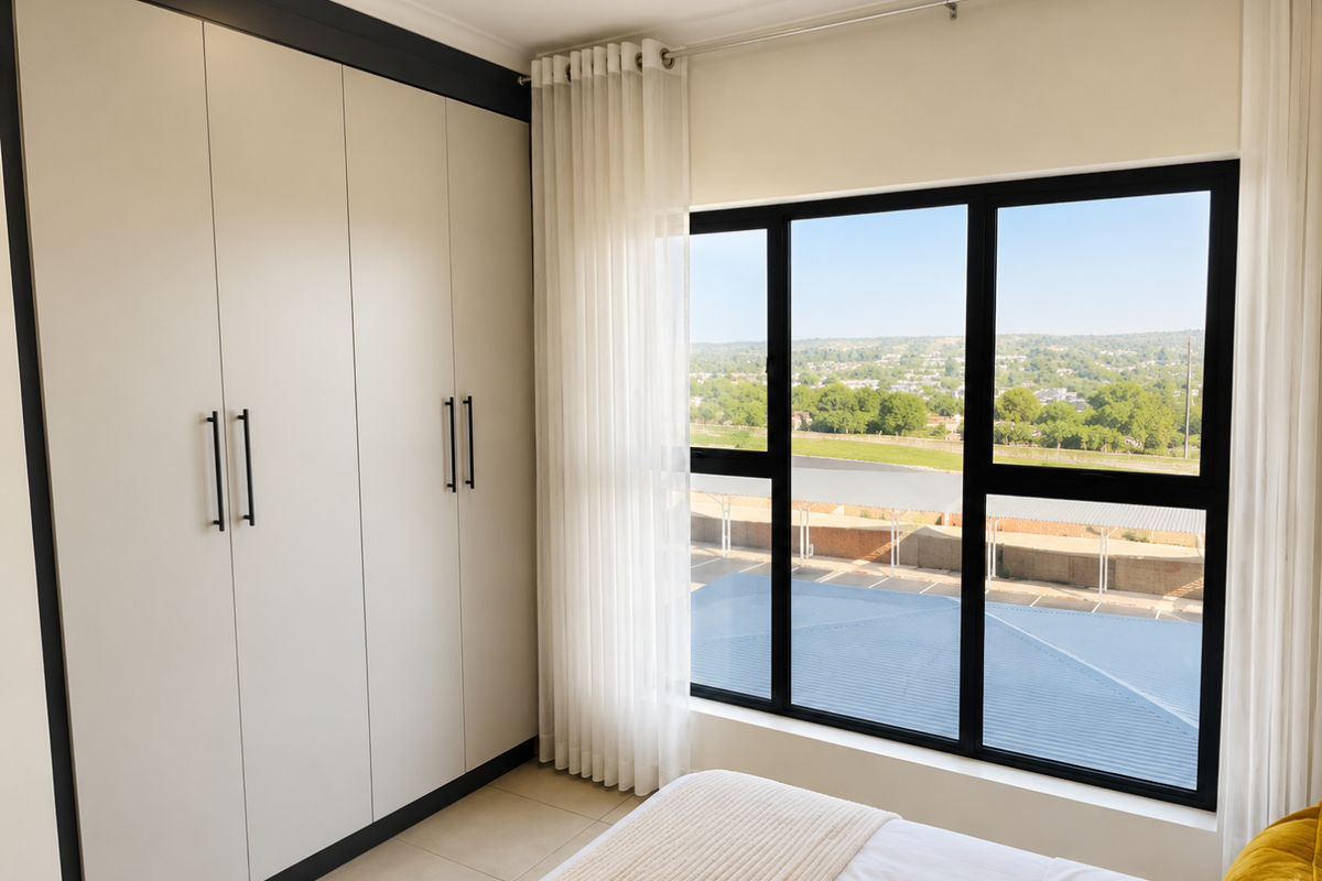 A bright bedroom featuring modern built-in cupboards with black trim and large black-framed windows offering an elevated view of the surrounding area.