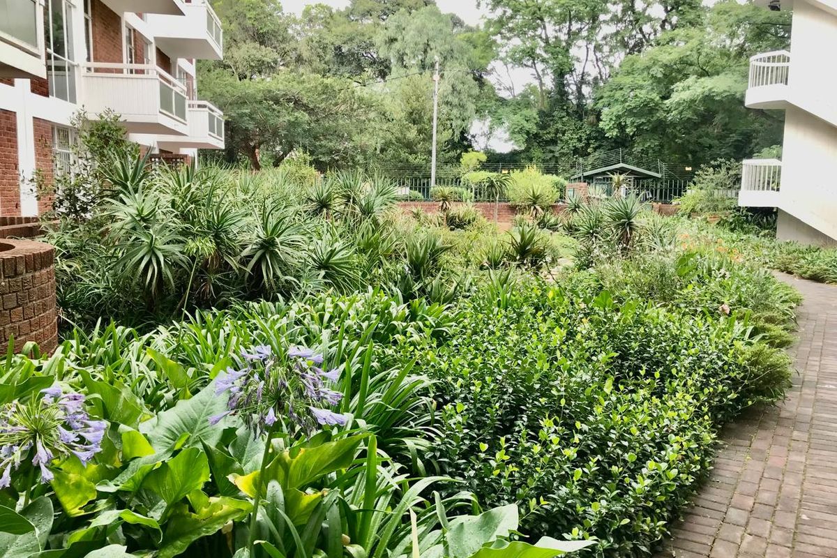 A view of the apartment complex rear garden area featuring established landscaping, brick paving, and private balconies overlooking the green space.