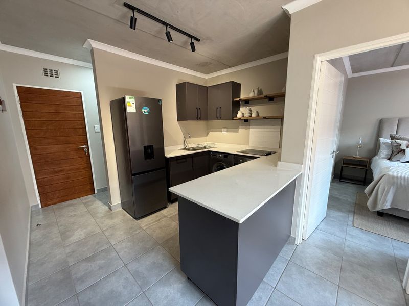 Modern kitchen featuring dark cabinetry, quartz countertops, a breakfast bar, and tiled flooring, with an open-plan layout connecting to the adjacent bedroom.