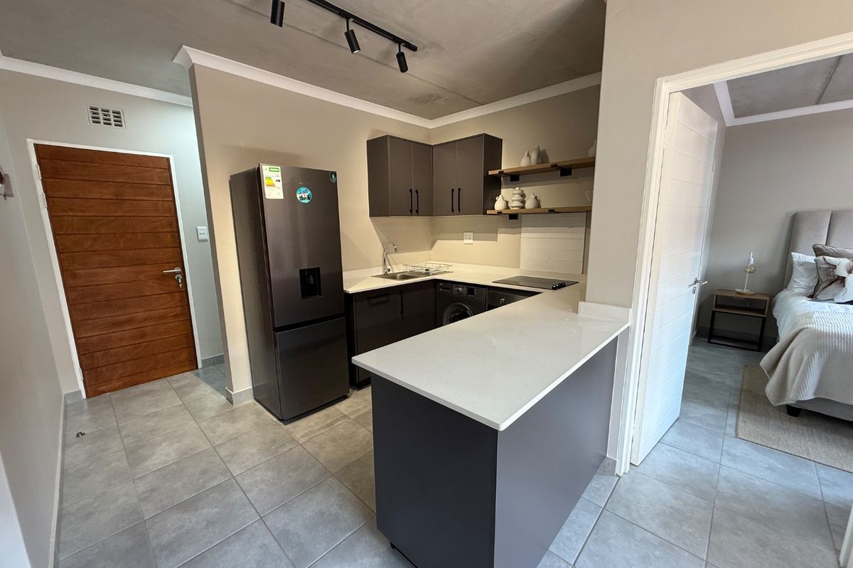 Modern kitchen featuring dark cabinetry, quartz countertops, a breakfast bar, and tiled flooring, with an open-plan layout connecting to the adjacent bedroom.