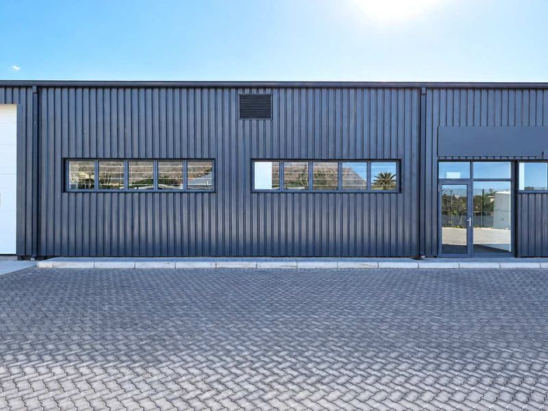Modern industrial warehouse facade featuring dark corrugated metal cladding, a large roller shutter door, glass entrance, and a clean paved forecourt.