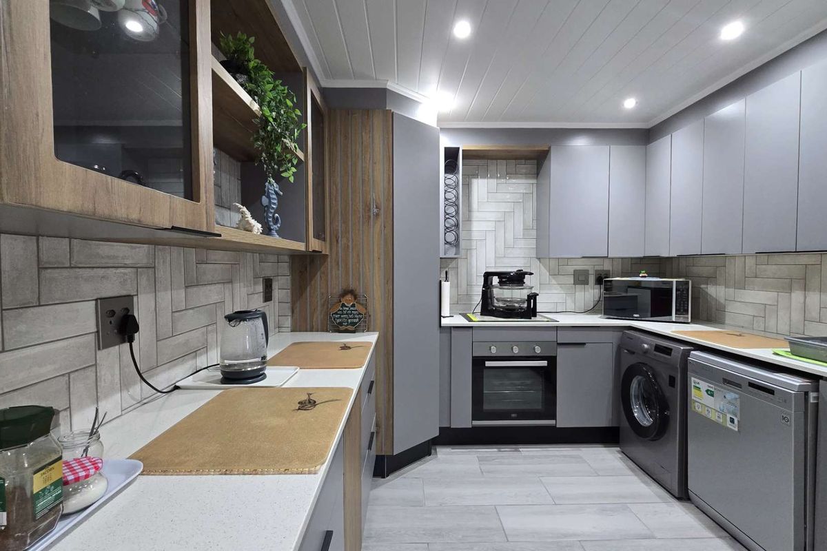 Modern kitchen featuring grey cabinetry, herringbone tiled backsplash, stone countertops, built-in oven, hob, and integrated space for laundry appliances.