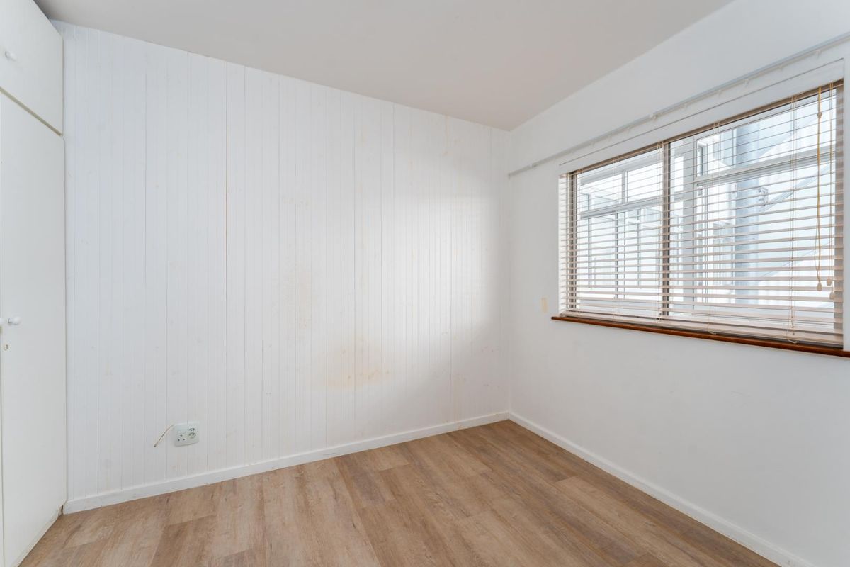 A bright, empty bedroom featuring wood-look flooring, white vertical wall paneling, a built-in cupboard, and a window with fitted blinds.
