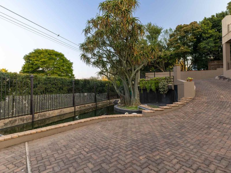 A paved driveway leading to the property entrance, featuring a landscaped water feature, mature tree, and secure perimeter fencing.