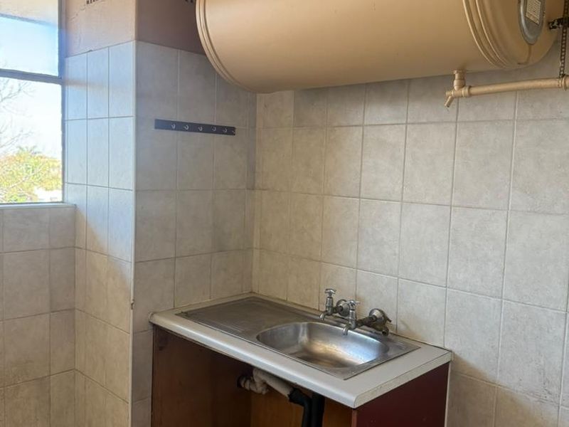 A functional laundry area featuring a stainless steel sink, wall-mounted electric water heater, and tiled finishes, currently requiring cosmetic updates and cabinet repairs.