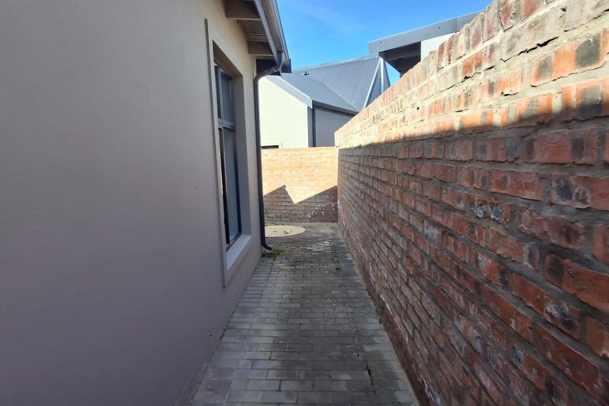 A narrow side passage featuring a paved walkway bordered by a high brick boundary wall and the exterior wall of the house.