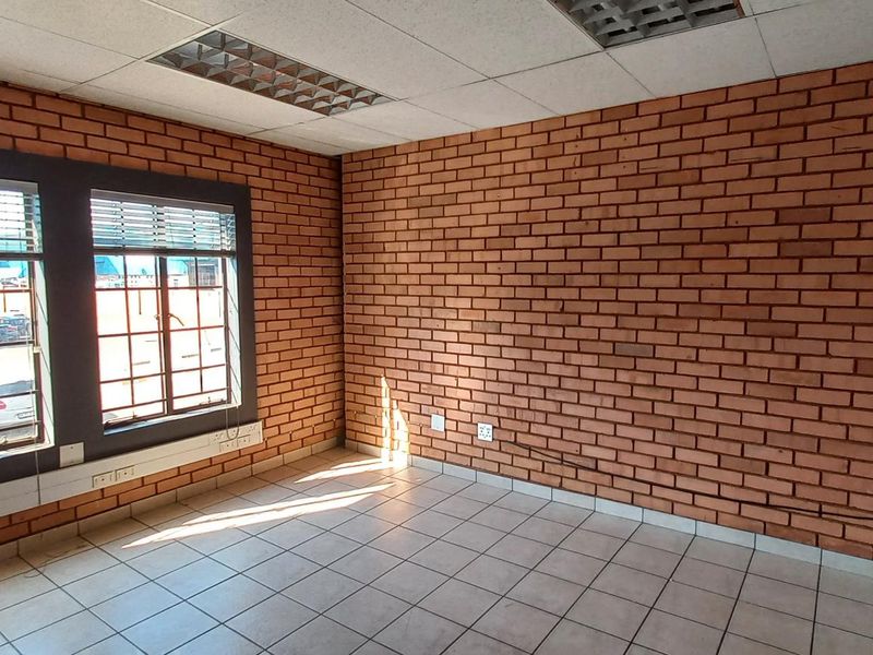 An empty office space featuring exposed brick walls, tiled flooring, and recessed ceiling lighting with large windows providing natural light.