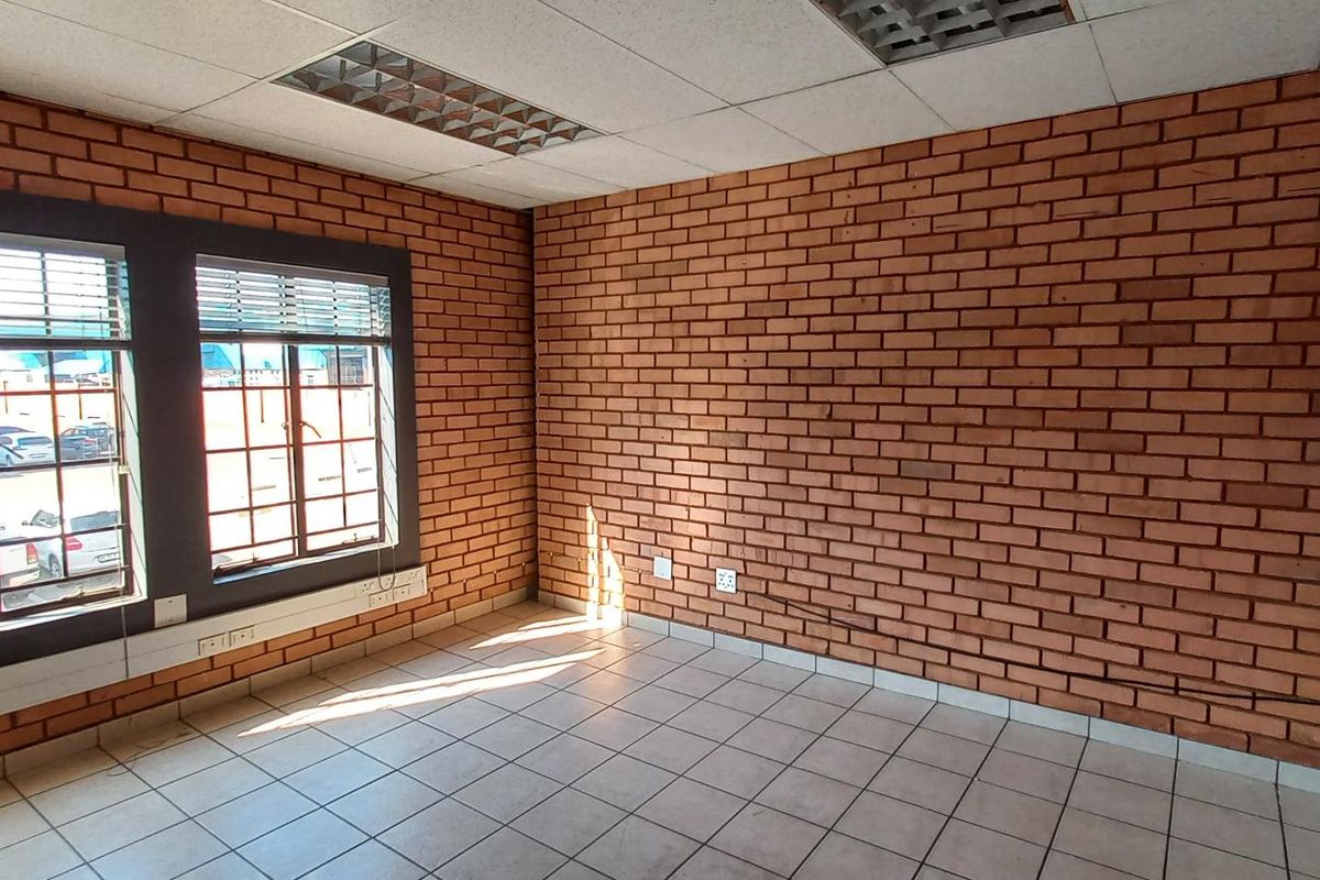 An empty office space featuring exposed brick walls, tiled flooring, and recessed ceiling lighting with large windows providing natural light.