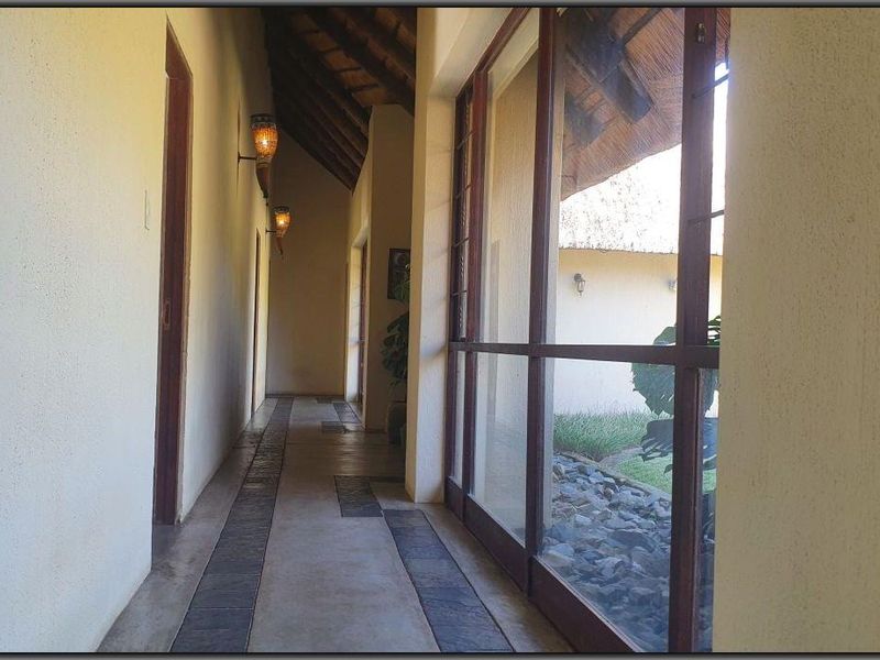 A bright, spacious hallway featuring a high thatched roof, stone tile flooring, and large glass windows overlooking an outdoor garden area.