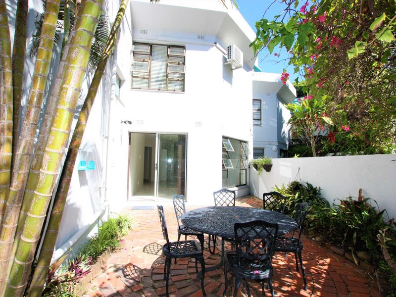 A private rear courtyard featuring brick paving, established garden beds, and direct access to the townhouse interior via a sliding glass door.