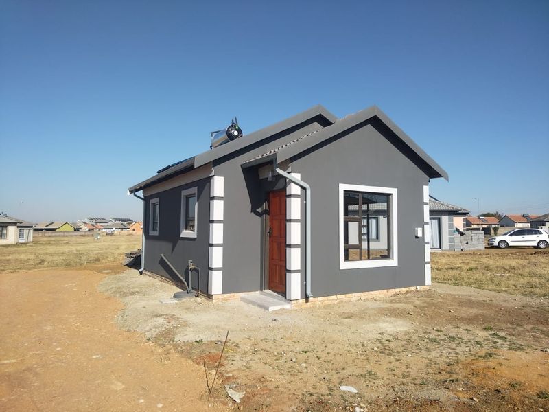 A modern, compact single-story house featuring a grey plastered facade, white corner detailing, and a solar water heating system on the roof.