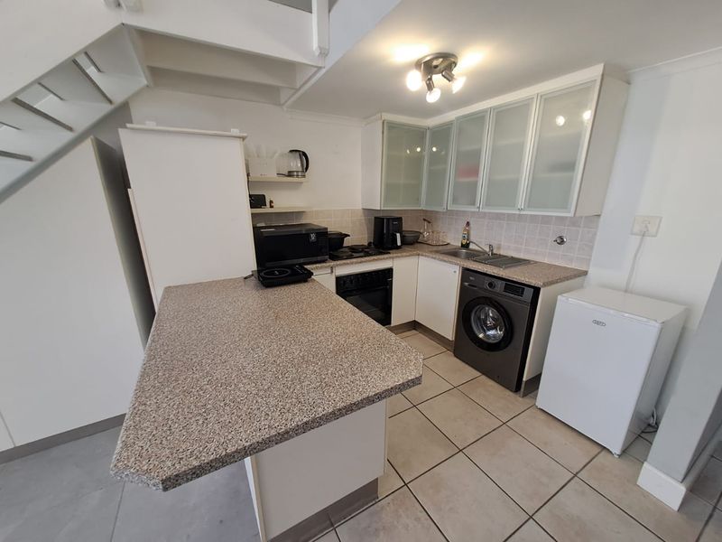 Compact kitchen featuring granite-look countertops, built-in oven, hob, and tiled flooring, conveniently located beneath a staircase in an open-plan apartment layout.