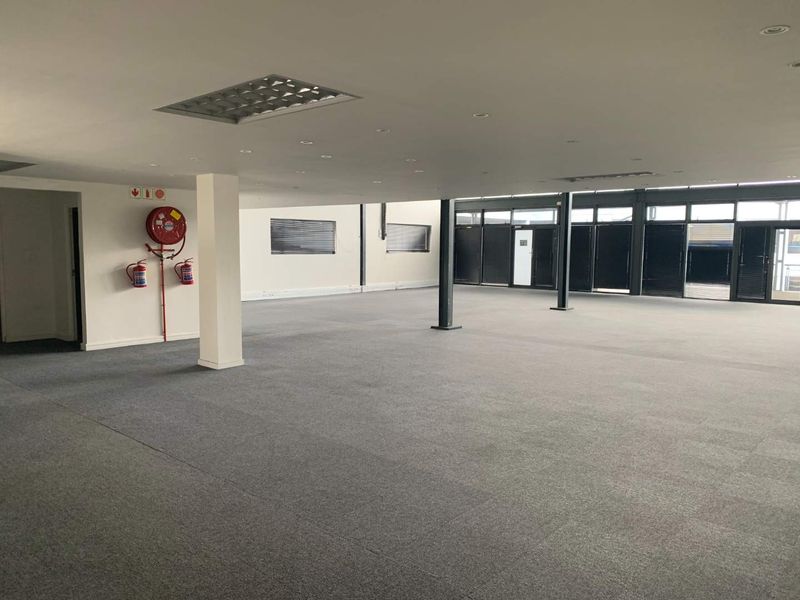A spacious, open-plan office area featuring carpeted flooring, structural columns, recessed lighting, and large windows with fitted blinds.