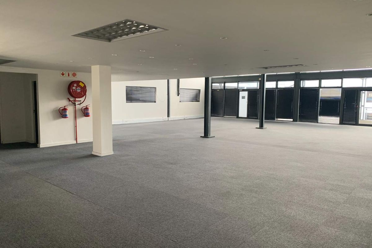 A spacious, open-plan office area featuring carpeted flooring, structural columns, recessed lighting, and large windows with fitted blinds.