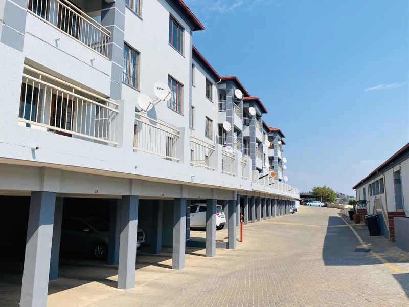 Exterior view of an apartment complex featuring covered ground-level parking bays, upper-level balconies, and a paved driveway.