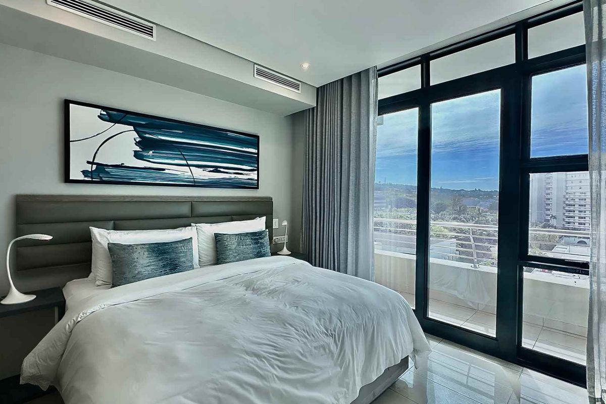 Modern bedroom featuring floor-to-ceiling windows with balcony access, ducted air conditioning, and sleek tiled flooring, offering a bright and contemporary living space.