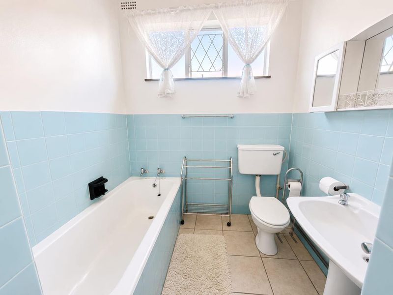 A functional bathroom featuring a bathtub, pedestal basin, and toilet with classic light blue wall tiling and a leaded glass window.