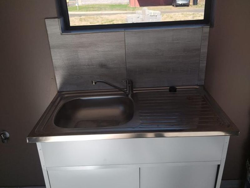 A compact kitchen sink unit featuring a stainless steel basin, mixer tap, and tiled splashback, positioned beneath a window.