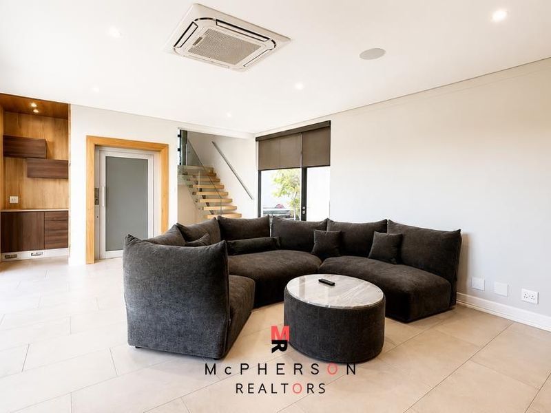 Modern open-plan living area featuring tiled floors, recessed lighting, cassette air conditioning, and a sleek staircase with glass balustrade leading to the upper level.