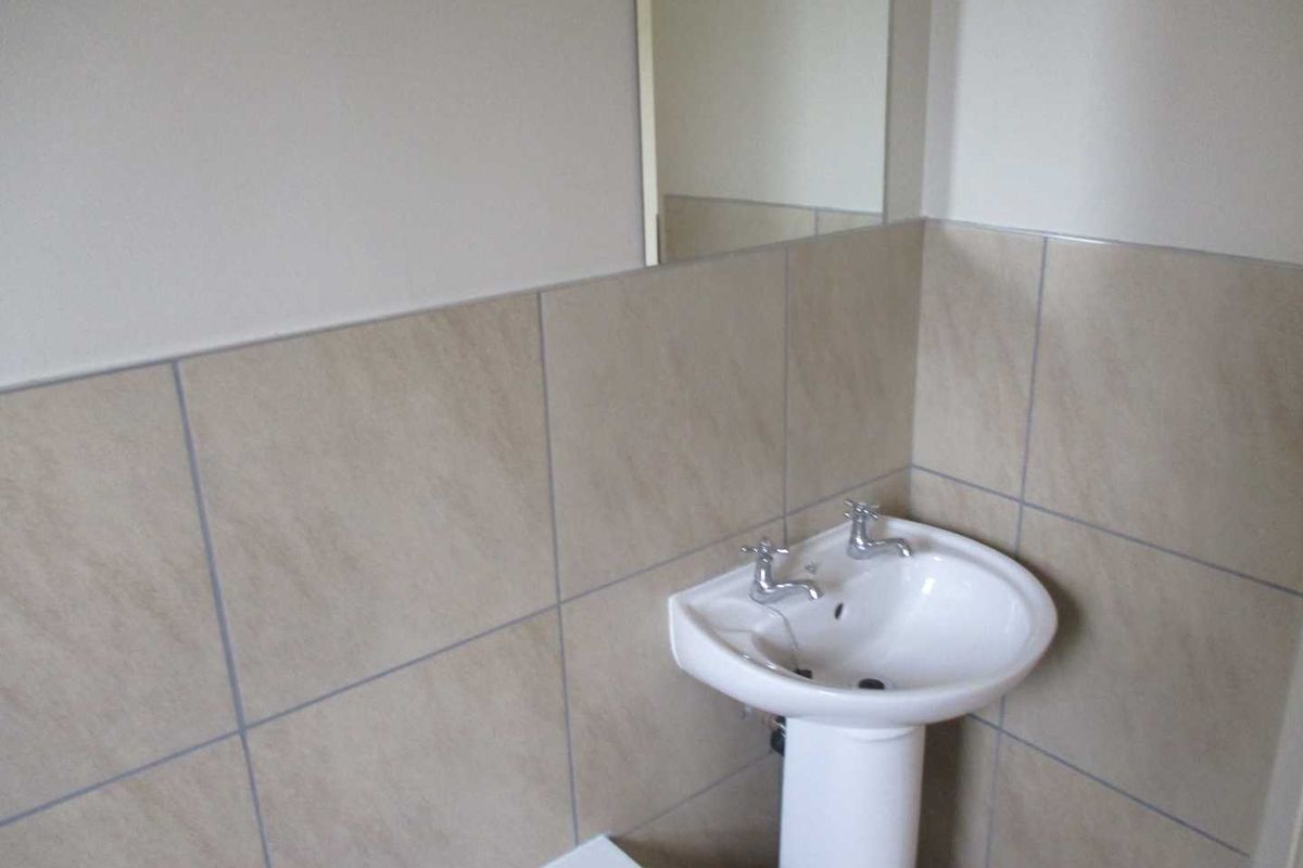 A clean, functional bathroom featuring a white pedestal basin, dual taps, and neutral ceramic wall tiling with an integrated mirror.