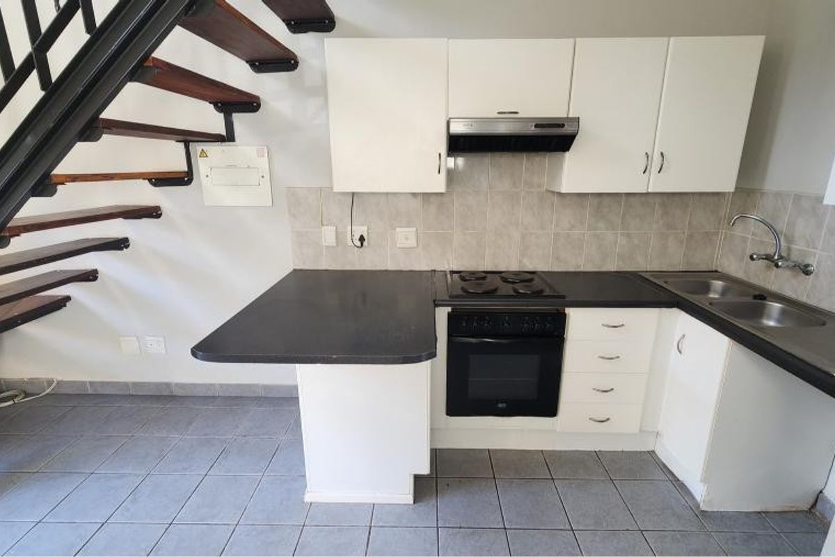 Compact kitchen featuring built-in cabinetry, electric hob, oven, and double sink, situated beneath an open-tread staircase with tiled flooring throughout.