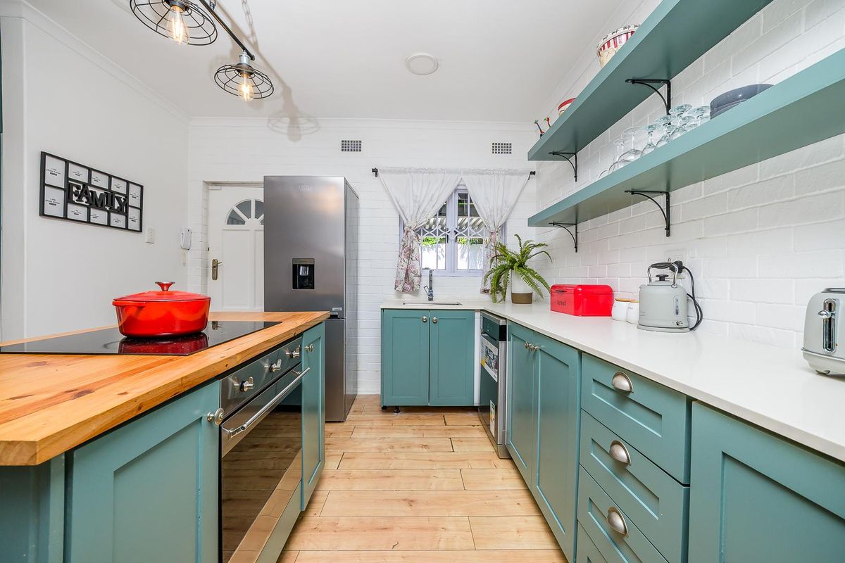 A well-appointed kitchen featuring teal cabinetry, timber countertops, a built-in oven with hob, and open shelving against a painted brick wall.