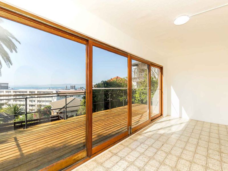 An empty sunroom or enclosed balcony featuring large sliding glass doors, wooden decking, and a scenic coastal view.