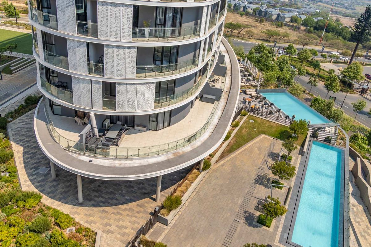 Aerial view of a modern apartment building featuring curved balconies, glass balustrades, and communal swimming pools set within landscaped grounds.