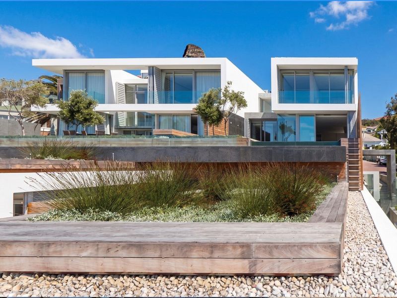 Modern multi-level townhouse exterior featuring extensive glass sliding doors, timber decking, and tiered landscaped garden areas with stone retaining walls.