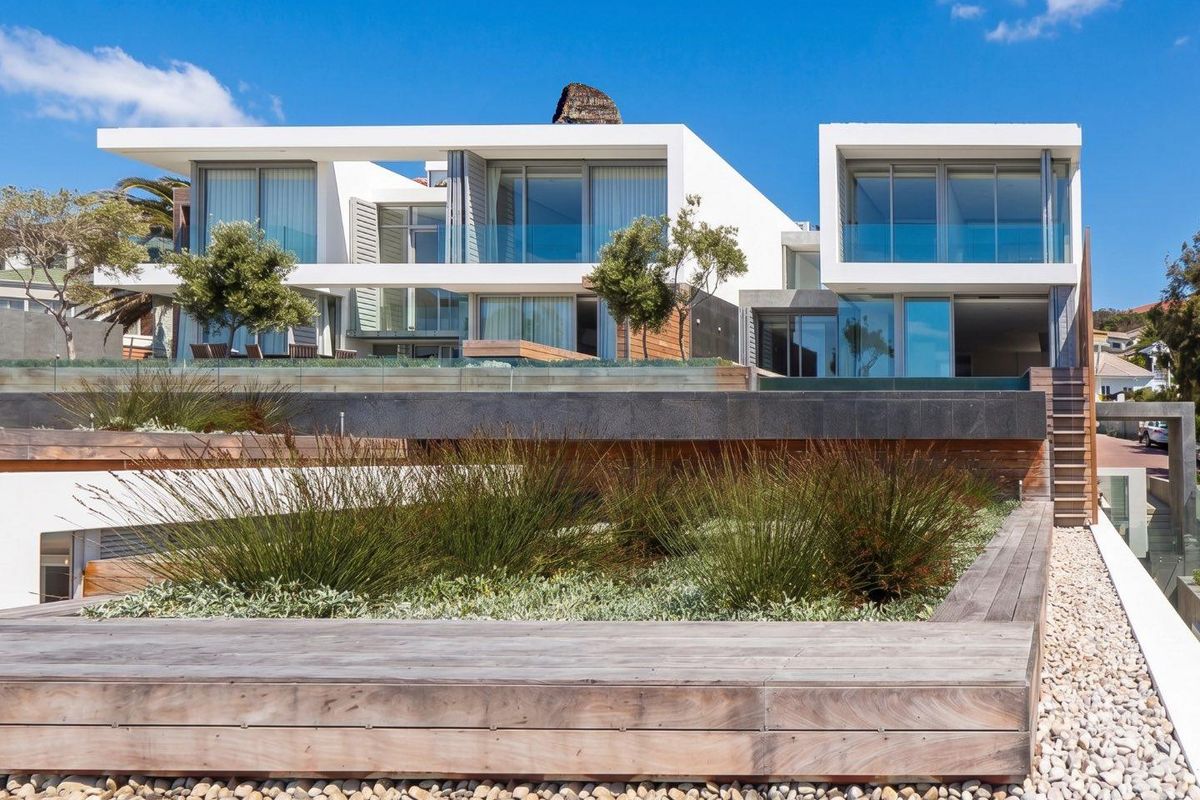 Modern multi-level townhouse exterior featuring extensive glass sliding doors, timber decking, and tiered landscaped garden areas with stone retaining walls.