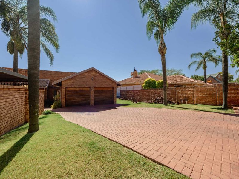 Front exterior view featuring a double garage, paved brick driveway, and established garden with palm trees and brick boundary walls.
