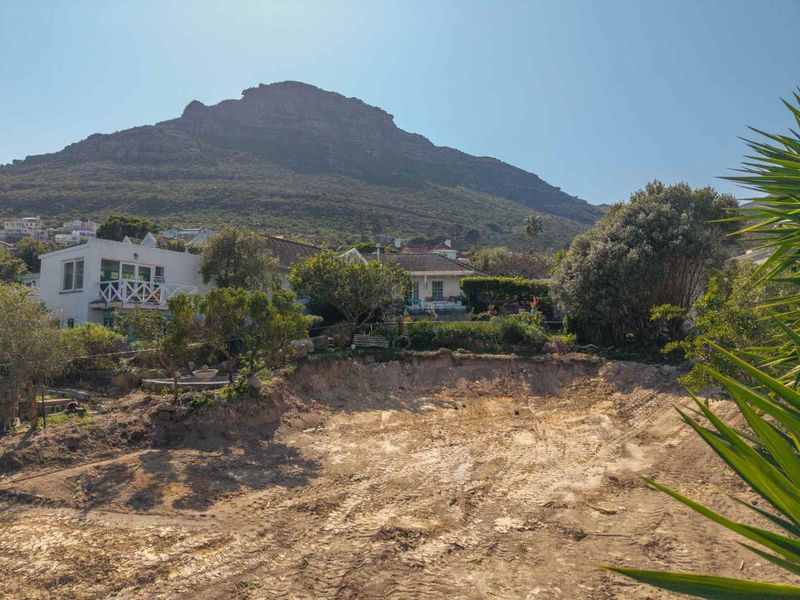 A view of a residential plot featuring significant earth excavation, mature garden landscaping, and a scenic mountain backdrop.
