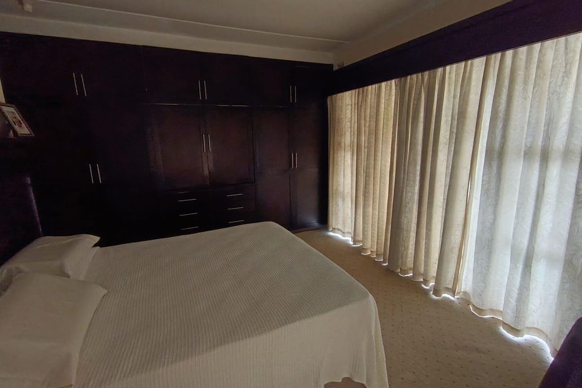 A spacious bedroom featuring extensive dark wood built-in cupboards, neutral carpeting, and large windows covered by floor-to-ceiling curtains.