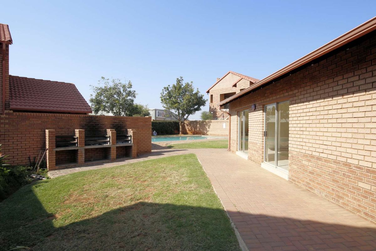 Rear exterior view featuring a paved patio, built-in brick braai area, lawn, and access to a communal swimming pool.