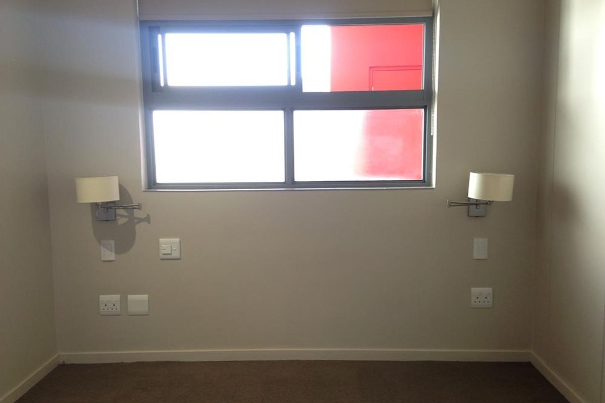 A clean, empty bedroom featuring neutral walls, carpeted flooring, aluminium window frames with a roller blind, and wall-mounted bedside lighting fixtures.