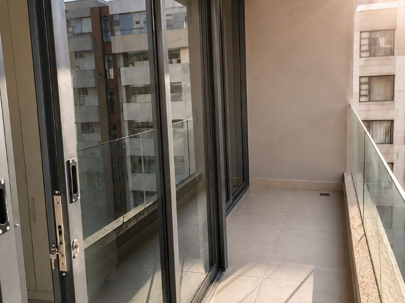A modern, covered balcony featuring sleek sliding glass doors, a contemporary glass balustrade, and neutral floor tiling with recessed lighting.